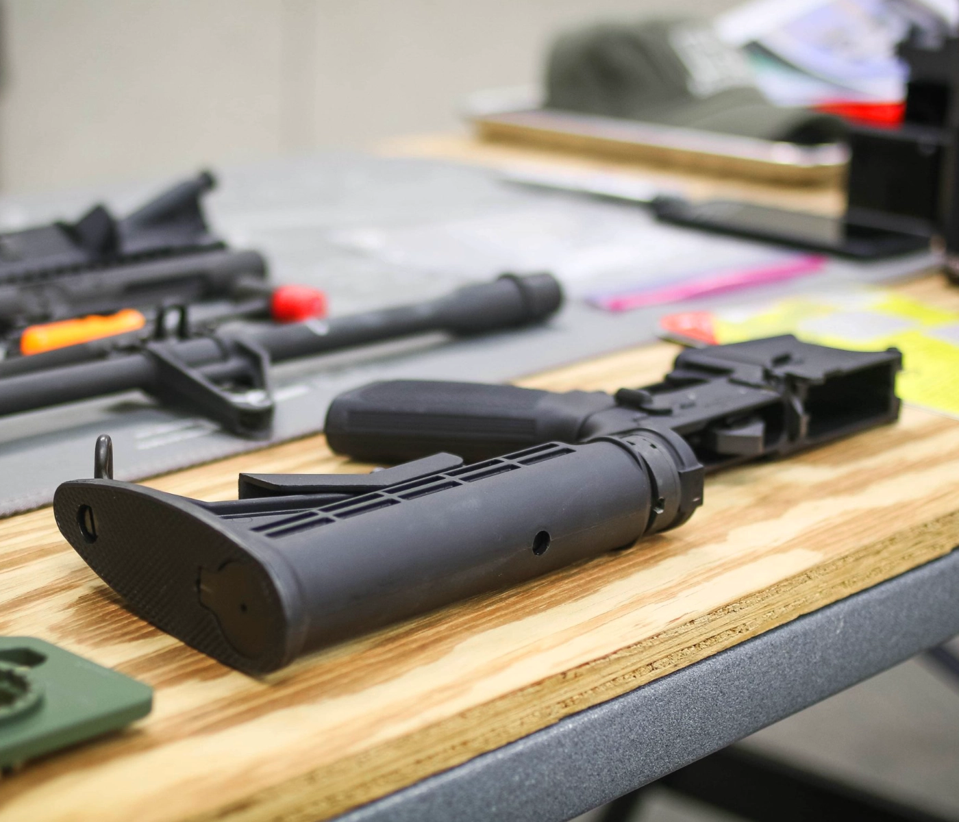 A close-up of a black rifle stock and lower receiver lying on a wooden table, with firearm components and tools blurred in the background at a POST AR armorer certification St. Louis course.