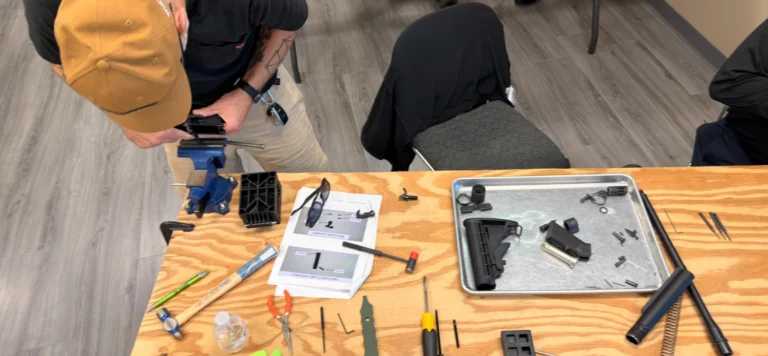 A person in a tan cap works on assembling or repairing a black device at a wooden table cluttered with tools, parts, and an instruction manual—perhaps practicing skills from an AR armorer course St. Louis. An empty chair is nearby; the floor is gray wood-patterned.