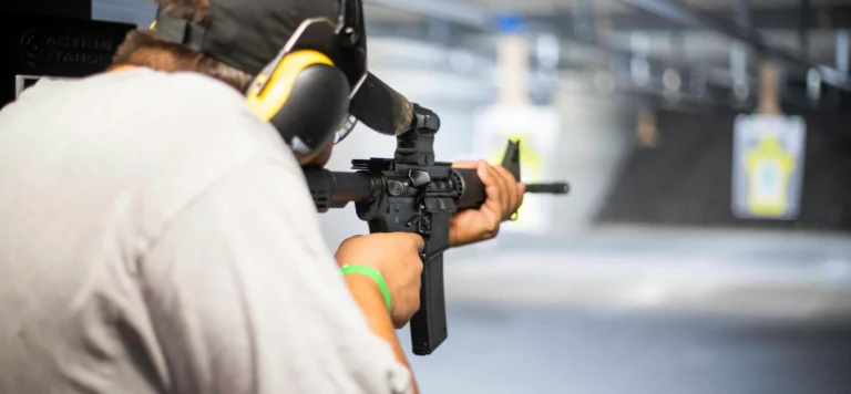 A person wearing ear protection aims a rifle at a paper target at an indoor shooting range during POST AR-15 qualification in St. Louis. The scene is slightly blurred, focusing on the shooter and the firearm.