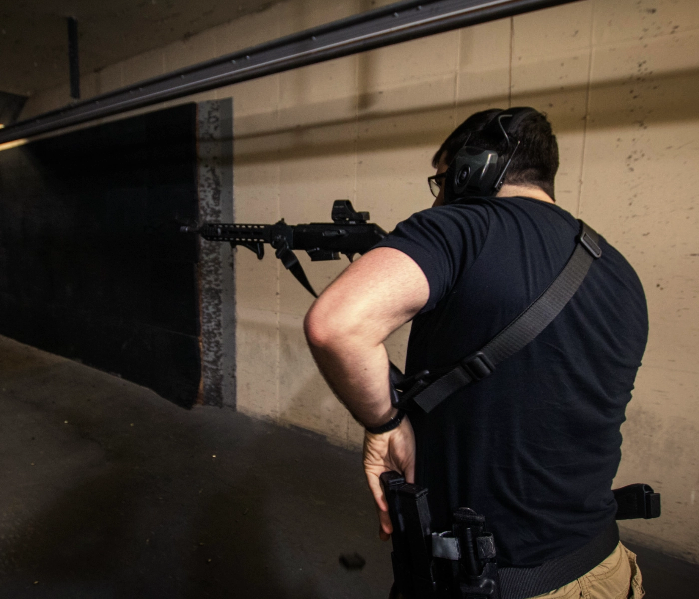A person wearing hearing protection and a black shirt stands at an indoor shooting range during a basic rifle course in St. Louis, aiming a rifle downrange. The concrete walls and target area are visible in the background.