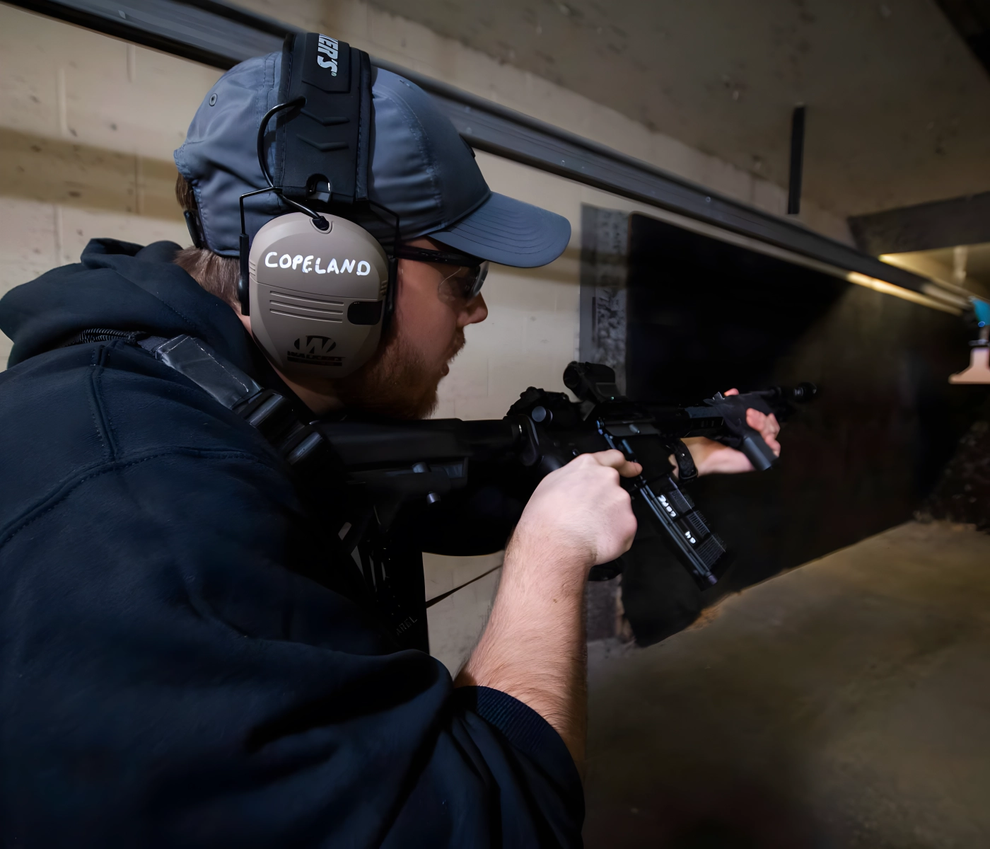 A person wearing ear protection, safety glasses, and a hat aims a rifle at an indoor shooting range during a basic rifle course in St. Louis. The individual appears focused, with both hands on the firearm.