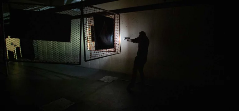 A person stands in a dimly lit room, aiming a handgun with a flashlight. Fenced partitions and targets hint at a low light shooting course in St. Louis, suggesting specialized training in challenging conditions.