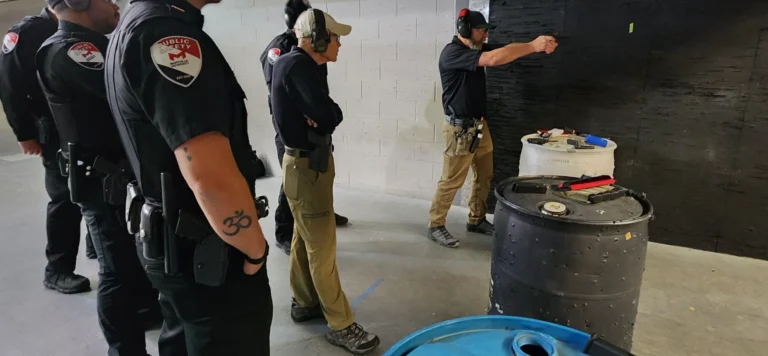 A group of people in uniforms stand in an indoor shooting range during a practical pistol course in St. Louis. One person aims a handgun at a target while others observe, with barrels and shooting accessories nearby.