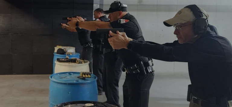 Four uniformed police officers practice aiming handguns at an indoor shooting range, standing in a line behind blue barrels with gear placed on top. They wear ear protection and safety glasses.