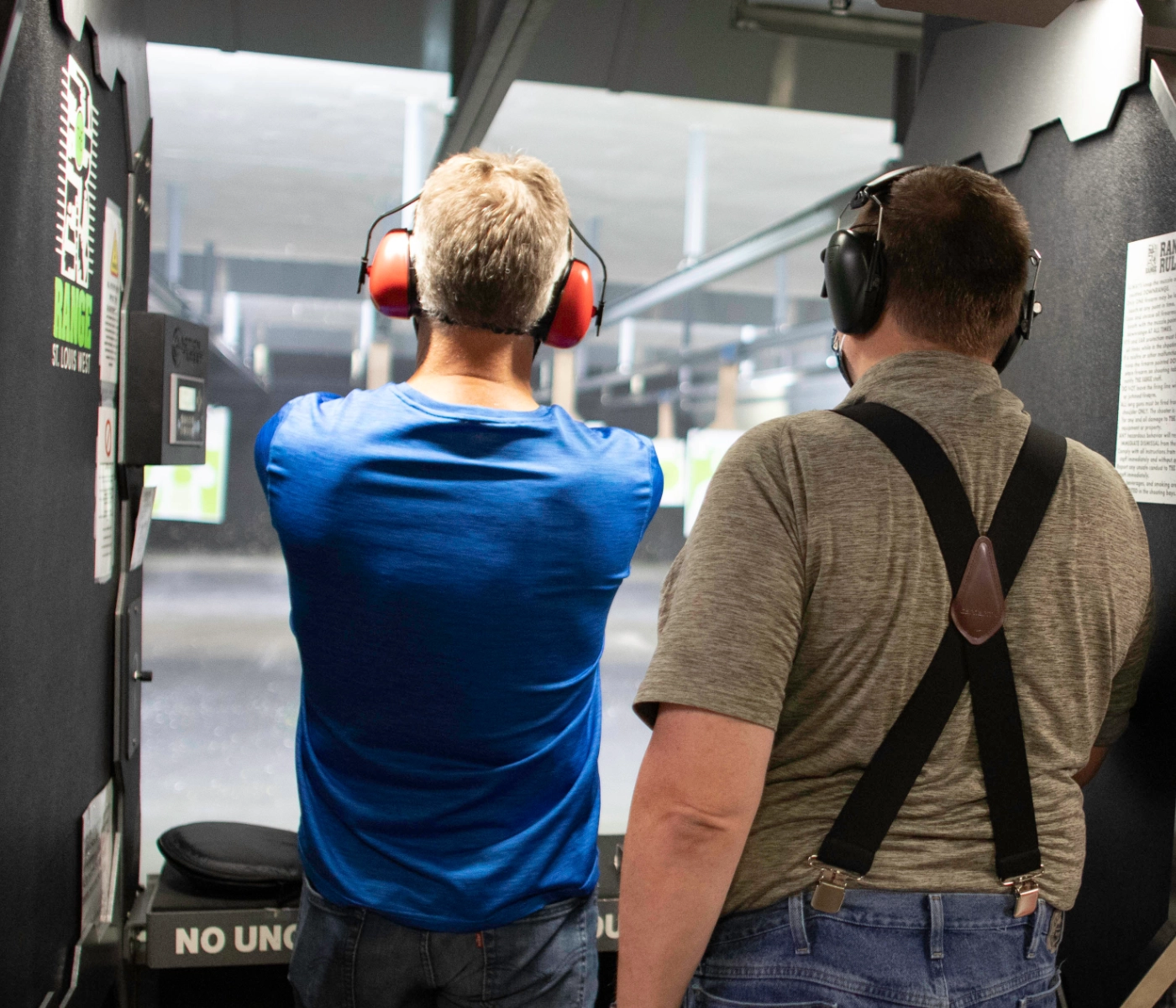 Two men wearing ear protection stand side by side at an indoor shooting range during an advanced red dot pistol course in St. Louis, aiming at paper targets downrange. The man on the left wears a blue shirt; the other sports a gray shirt with suspenders.