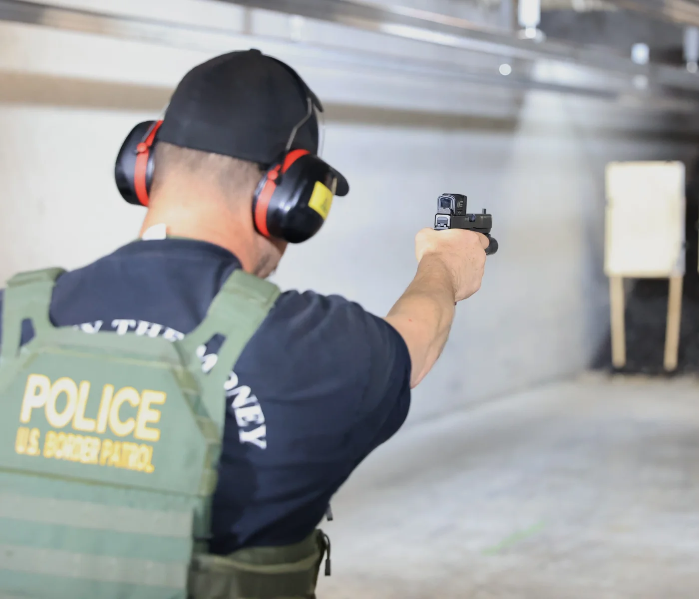 A U.S. Border Patrol officer wearing ear protection and a vest labeled "POLICE" aims a handgun during POST red dot qualification at a target downrange inside an indoor shooting range in St. Louis.