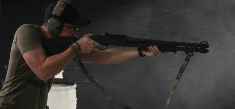 A person wearing ear protection and a cap aims a shotgun indoors at a shooting range, standing against a dark wall background—ideal for an intermediate shotgun course in St. Louis.
