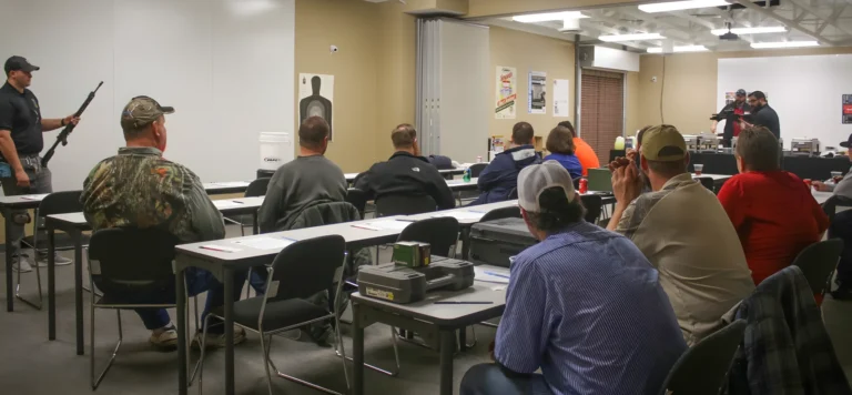 A group of people sit at desks in a classroom, facing two instructors at the front. One instructor holds a rifle while standing near a target poster, demonstrating shooting classes St. Louis to the class.