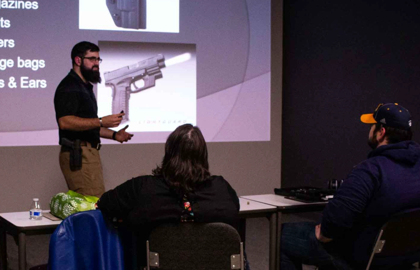 A bearded man stands and gives a new gun owner maintenance class presentation to two seated people in a St. Louis classroom, with a slide showing a handgun projected on the screen behind him.