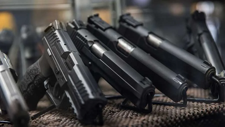 A row of black handguns is displayed on a counter, resting on a textured surface, in what appears to be a gun shop or display case.