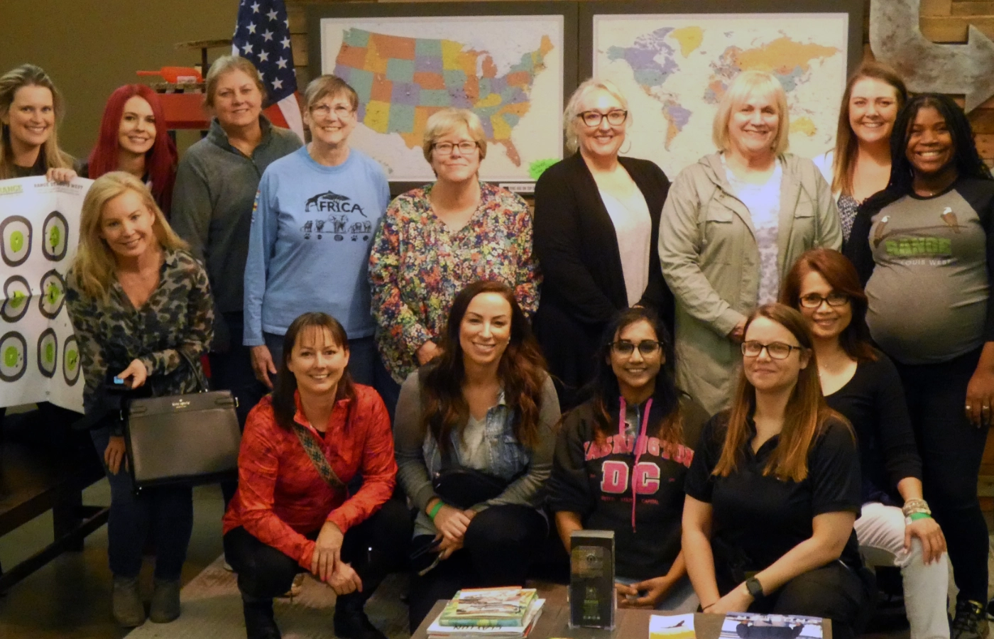 A group of fifteen women pose and smile together indoors during a women's self defense seminar in St. Louis, with maps of the United States and the world and an American flag visible in the background.