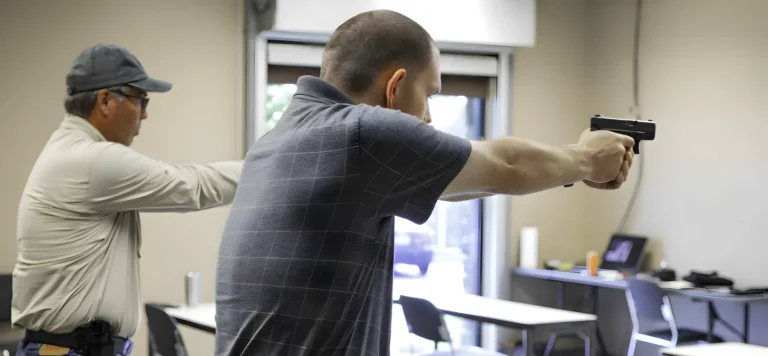 Two men standing indoors, each aiming a handgun towards the same direction, in what appears to be a firearms training or safety class; tables and chairs are visible in the background.